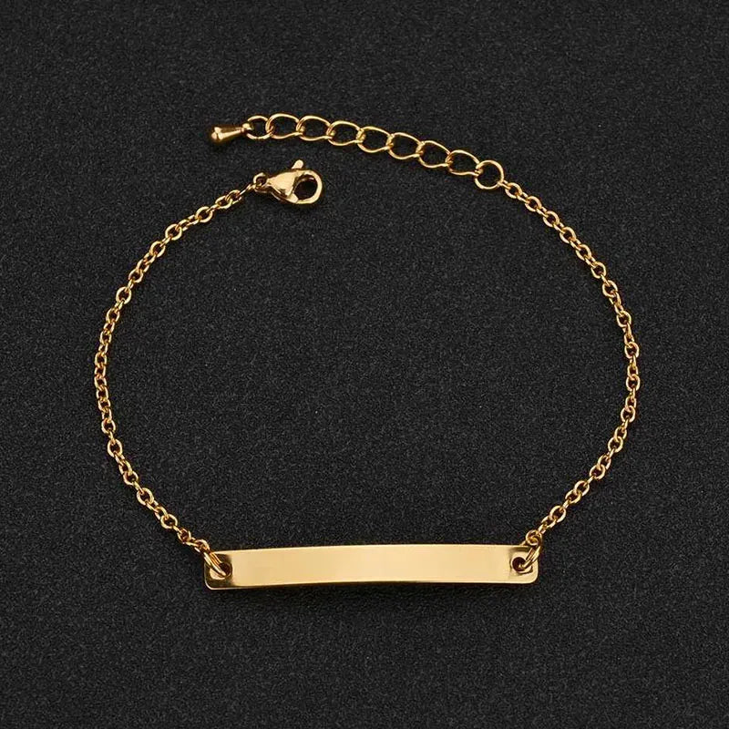 Gold thin chain bracelet with a plain rectangular nameplate and extension chain, on a black textured background.