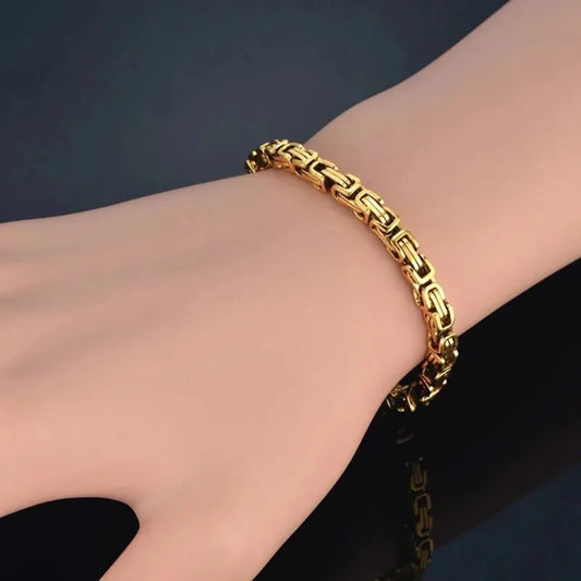 Gold Byzantine box-chain bracelet worn on the wrist, close-up against a dark background with reflection below.
