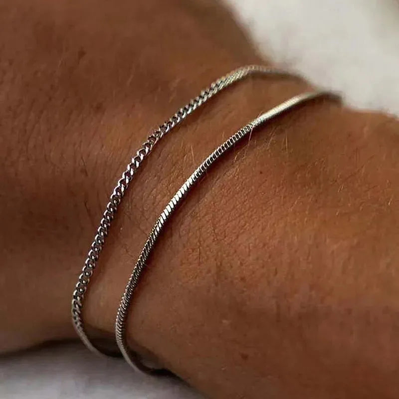 Two silver-tone chains layered on the wrist, close-up against a soft cream-colored background.