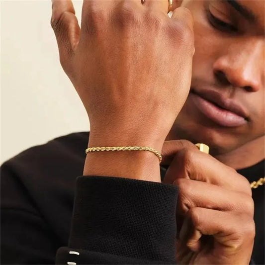 Gold twisted rope chain bracelet worn on the wrist, close to the face of a man in a black sweater with gold rings on his other hand.