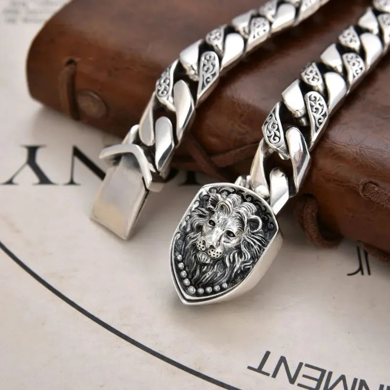 Close-up of the silver Cuban-link bracelet shown open, showcasing the sculpted lion-head shield plate, resting on a leather journal with a printed page in the background.