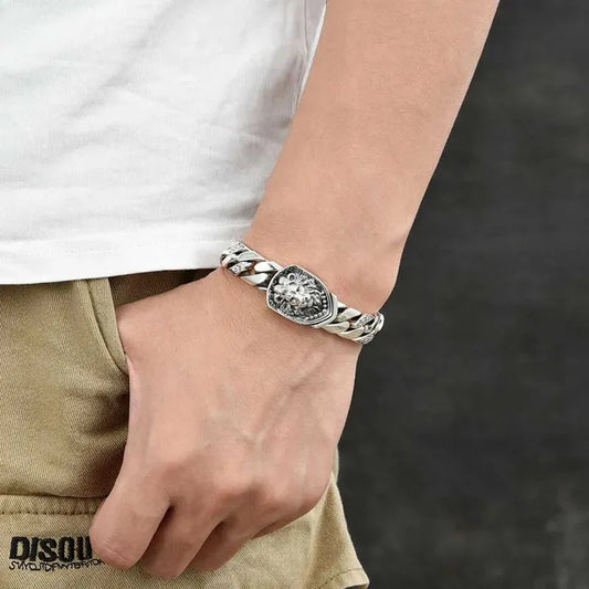 Silver Cuban-link bracelet with a lion-head shield centerpiece worn on the wrist, paired with a casual white t-shirt and khaki pants.