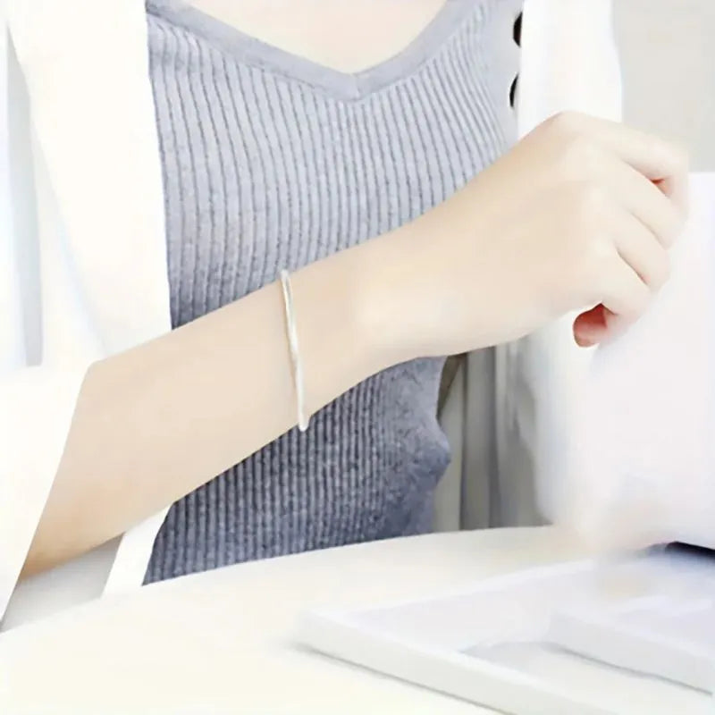 Silver snake chain bracelet worn on the wrist of a woman in a grey ribbed top and white cardigan, seated at a desk.