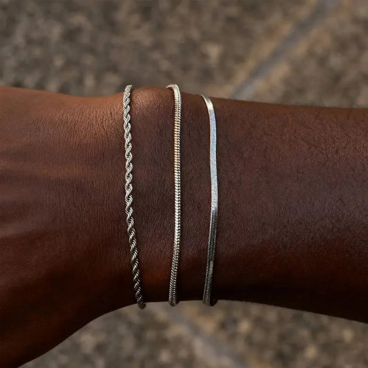 Three silver bracelets — a twisted rope chain, a snake chain, and a flat blade chain — stacked on the wrist, close-up against a stone tile background.