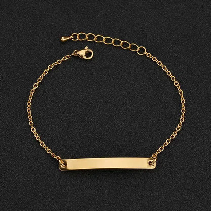 Gold thin chain bracelet with a plain rectangular nameplate and extension chain, on a black textured background.