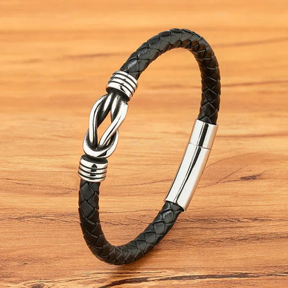 Black braided leather bracelet with a polished stainless steel infinity-knot clasp, on a wood background