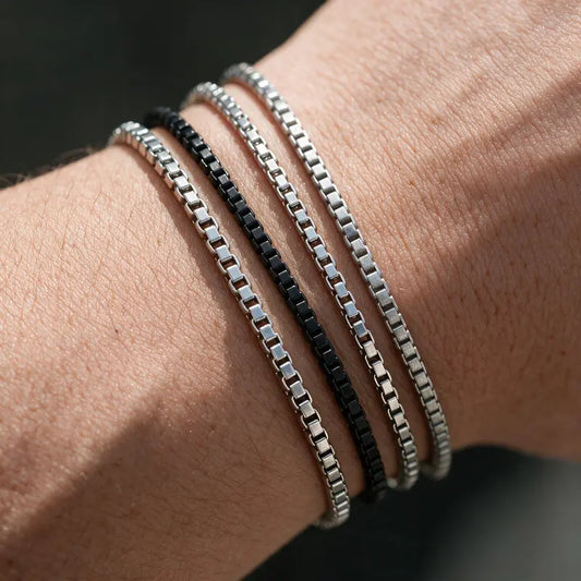 Four stacked box chain bracelets, one black and three silver, worn on the wrist