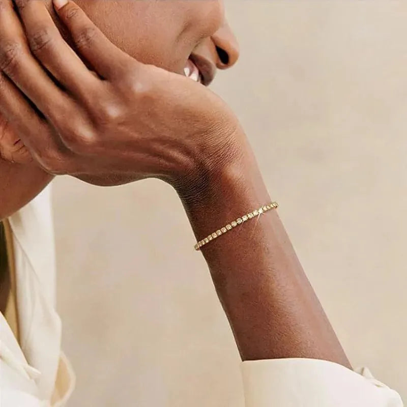 Gold tennis bracelet worn on the wrist of a person resting their hand against their cheek, cream-toned background.