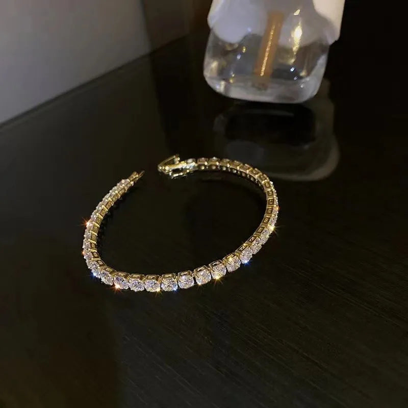 Gold tennis bracelet with round cubic zirconia stones laid in an oval on a dark wood surface beside a perfume bottle.