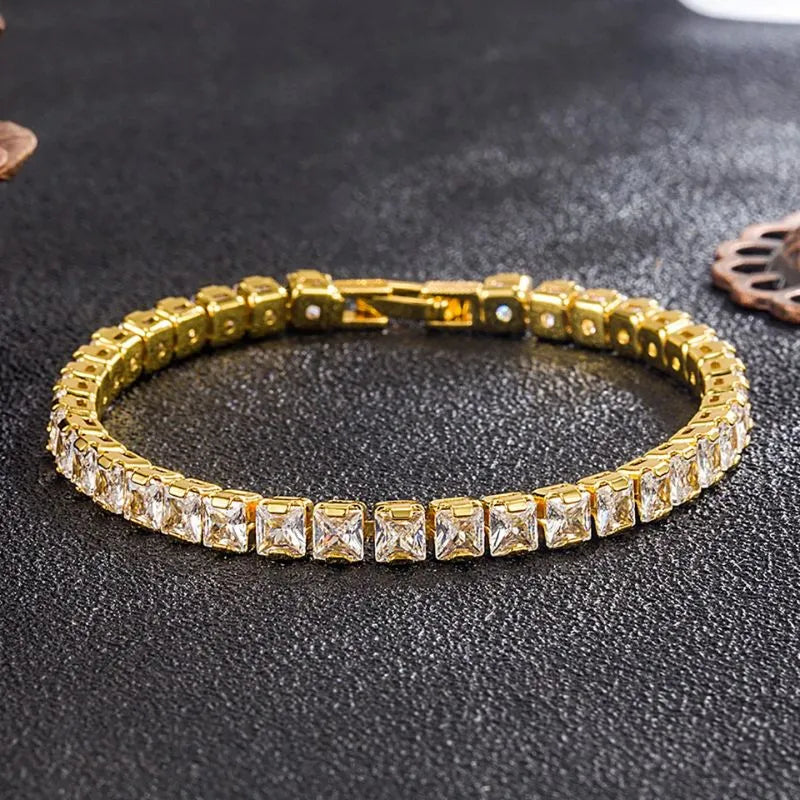 Gold tennis bracelet with square-cut cubic zirconia stones laid in a circle on a dark textured surface, with a decorative rustic ornament in the background.