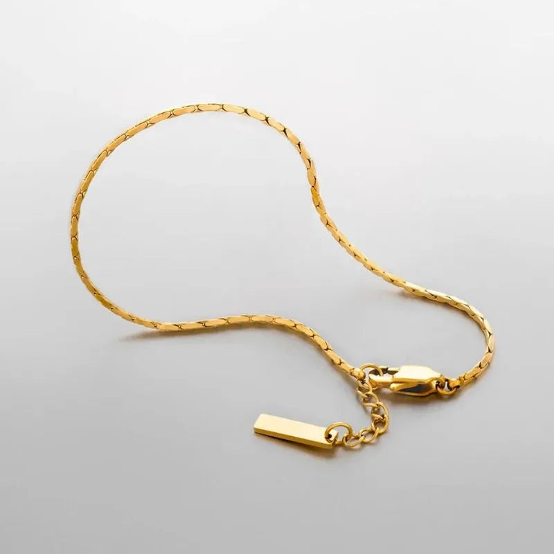 Gold wire chain bracelet draped in a loose curve on a light grey background, showing the clasp, bar tag charm, and extension chain.
