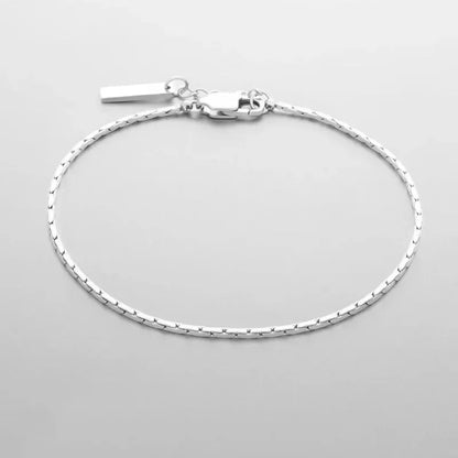 Silver-tone wire chain bracelet with a rectangular bar tag charm and lobster clasp, laid in an oval on a light grey background.