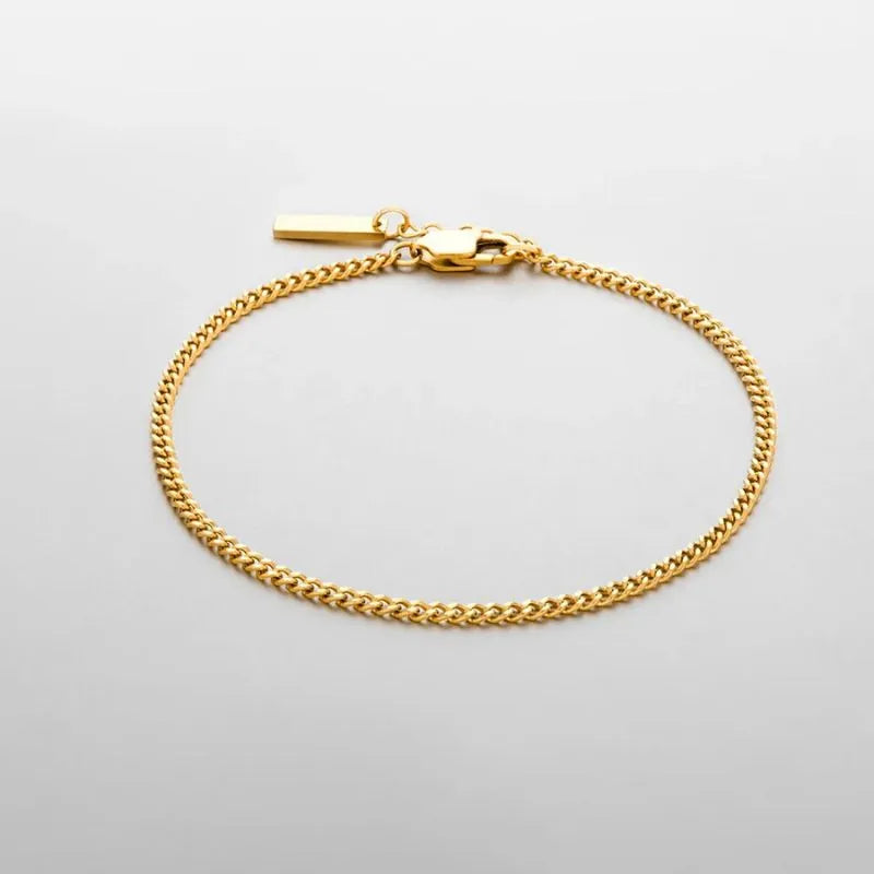 Thin gold Cuban curb chain bracelet with a small rectangular bar tag charm, on a plain white background