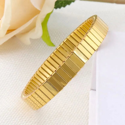 Gold stretch bracelet laid on white textured fabric beside a pale pink rose