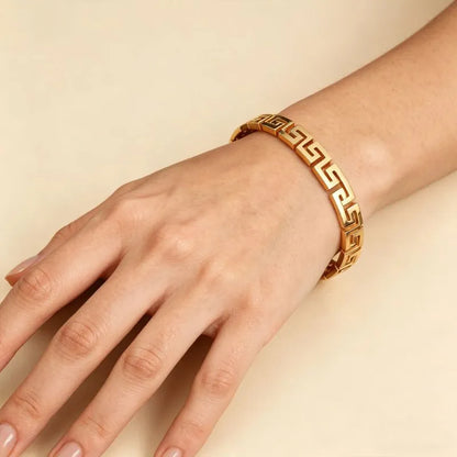 Close-up of a hand and wrist wearing the gold Greek key bangle bracelet, resting flat on a beige background