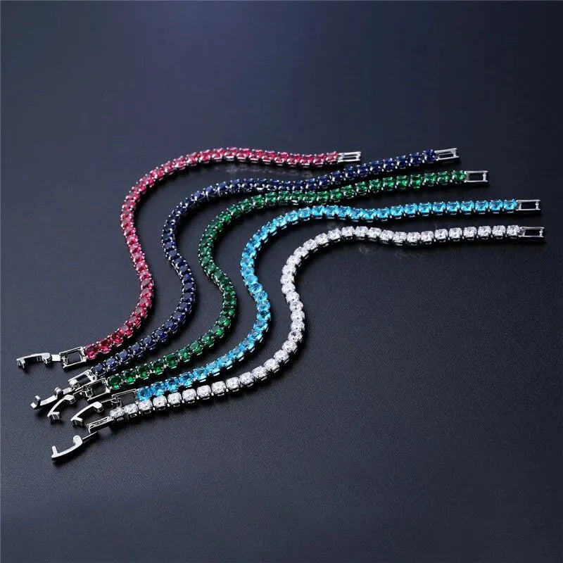 Five tennis bracelets in different coloured stones (pink, navy, green, light blue, clear) laid on a dark surface