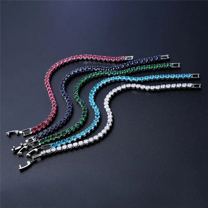 Five tennis bracelets in different coloured stones (pink, navy, green, light blue, clear) laid on a dark surface