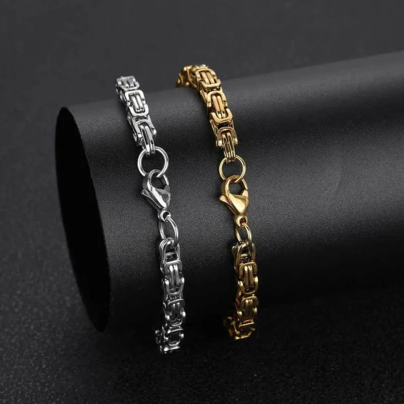 Silver and gold Byzantine box-chain bracelets displayed side by side, draped over a black cylindrical stand for a color comparison.
