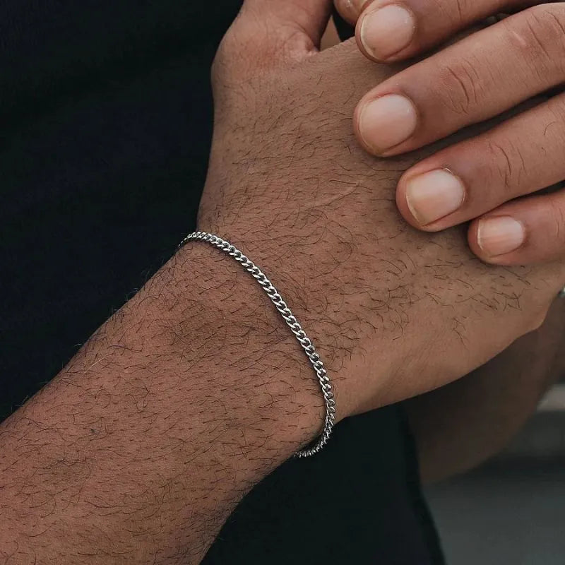 Thin silver-tone Cuban chain bracelet worn on the wrist, with the other hand's fingers resting on top against a black background.