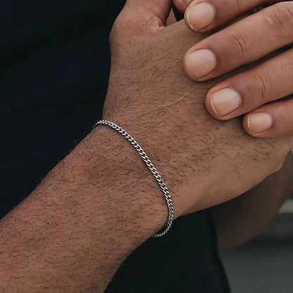 Thin silver-tone Cuban chain bracelet worn on the wrist, with the other hand's fingers resting on top against a black background.