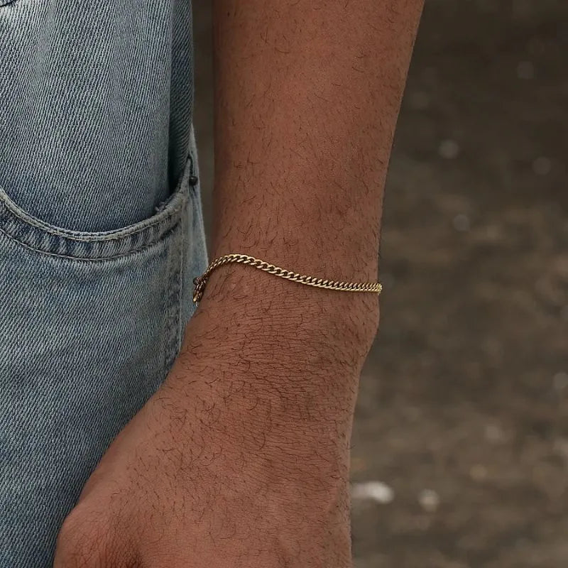 On-wrist gold Cuban chain bracelet shown outdoors with denim overalls in the background.