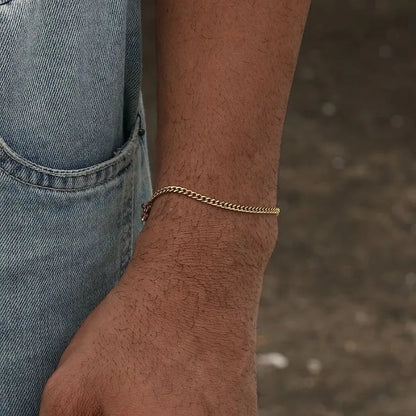 On-wrist gold Cuban chain bracelet shown outdoors with denim overalls in the background.