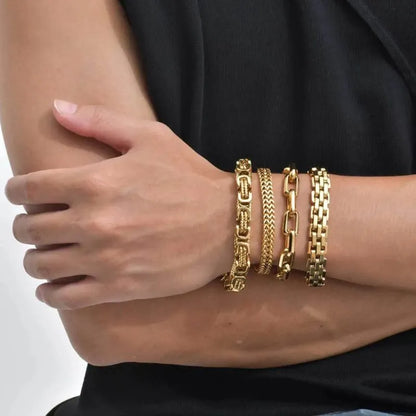Four gold chain bracelets stacked on a wrist with arms crossed, person wearing a black t-shirt