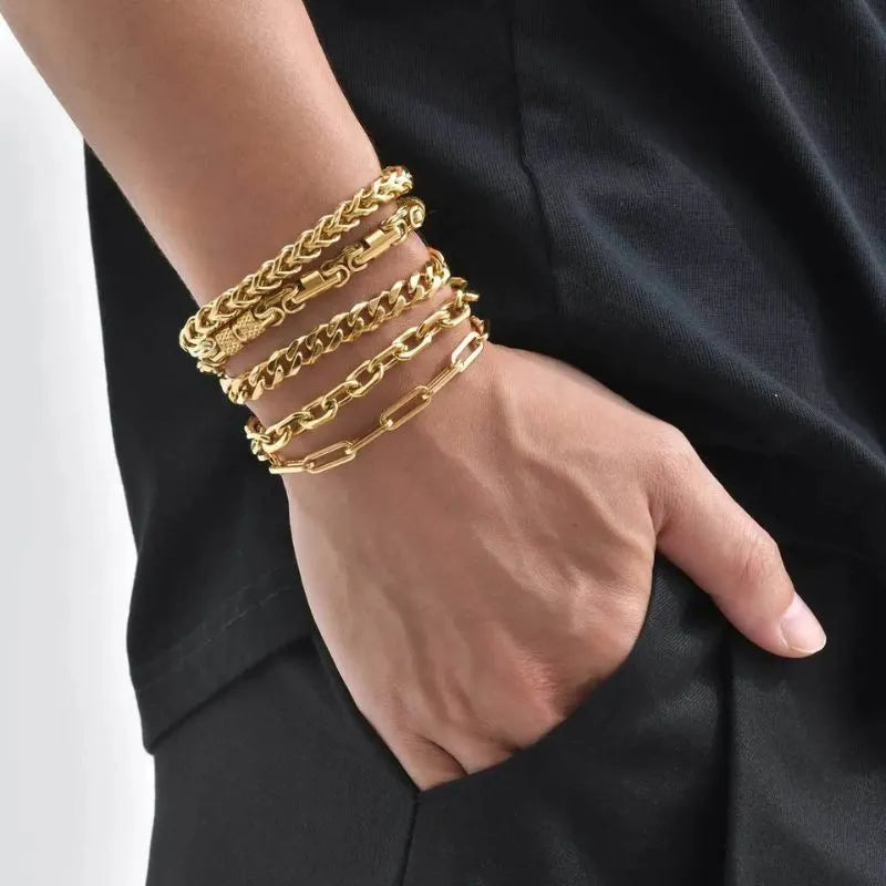 Wheat, curb and paperclip gold chain bracelets stacked on a wrist with hand in a trouser pocket