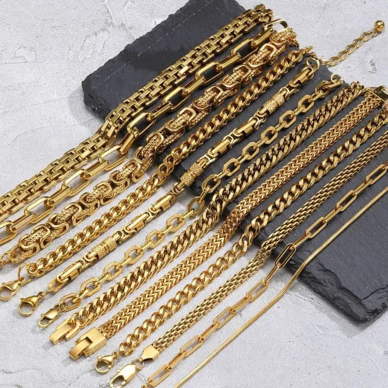 Close-up spread of about ten gold chain bracelet styles laid over a dark stone slab on a grey marble surface