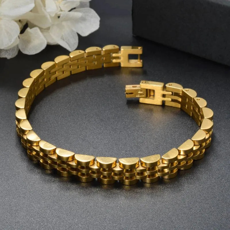 Gold watch link bracelet laid open in a circle on a dark textured surface with white flowers and a dark box