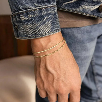 Two gold chains layered on the wrist, paired with a denim jacket sleeve in a casual indoor setting.