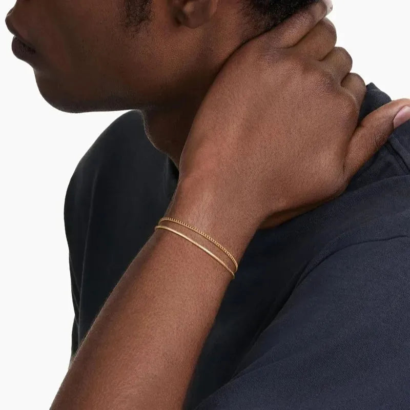 Two gold chains layered on the wrist of a man touching the back of his neck, wearing a dark grey t-shirt.
