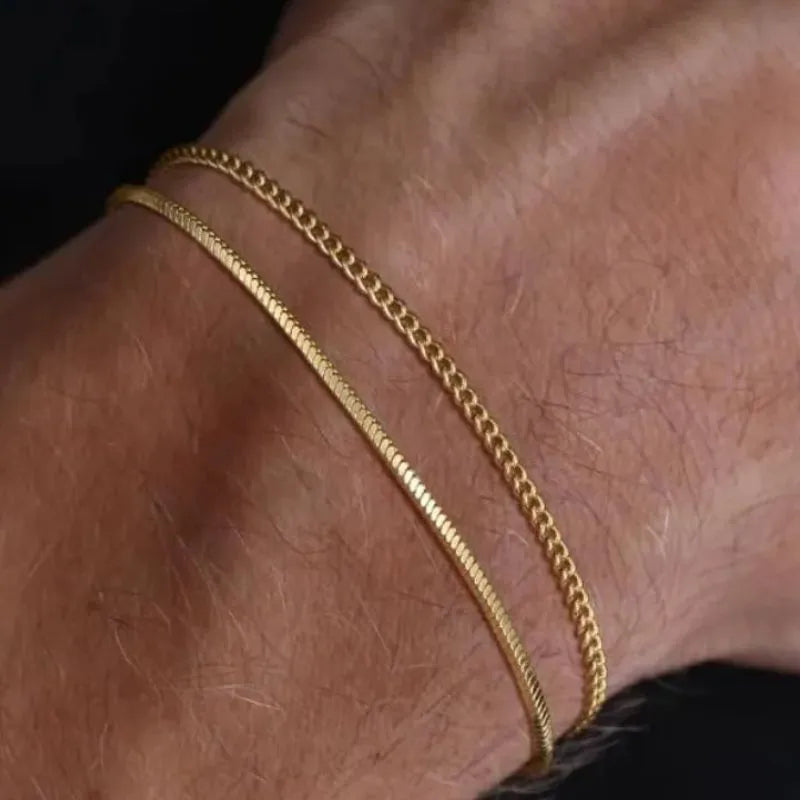 Two gold chains, a thin snake chain and a thicker Cuban-style chain, layered on the wrist, close-up against a black background.