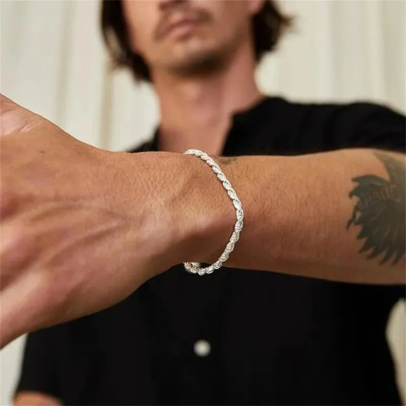 Silver-tone twisted rope chain bracelet worn on an extended wrist, with a man in a black shirt and forearm tattoo visible in the blurred background.