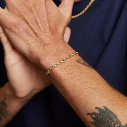 Gold twisted rope chain bracelet worn on the wrist with hands crossed, man in a navy shirt with a gold necklace and forearm tattoos visible.