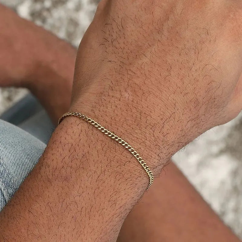 Gold Cuban link bracelet worn on the wrist outdoors, paired with denim shorts against a natural stone backdrop.