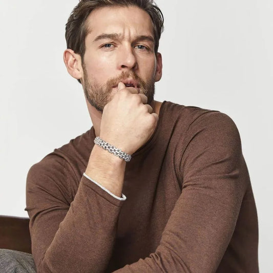 Silver watchband-link bracelet worn on the wrist of a man in a brown sweater, resting his hand near his chin.