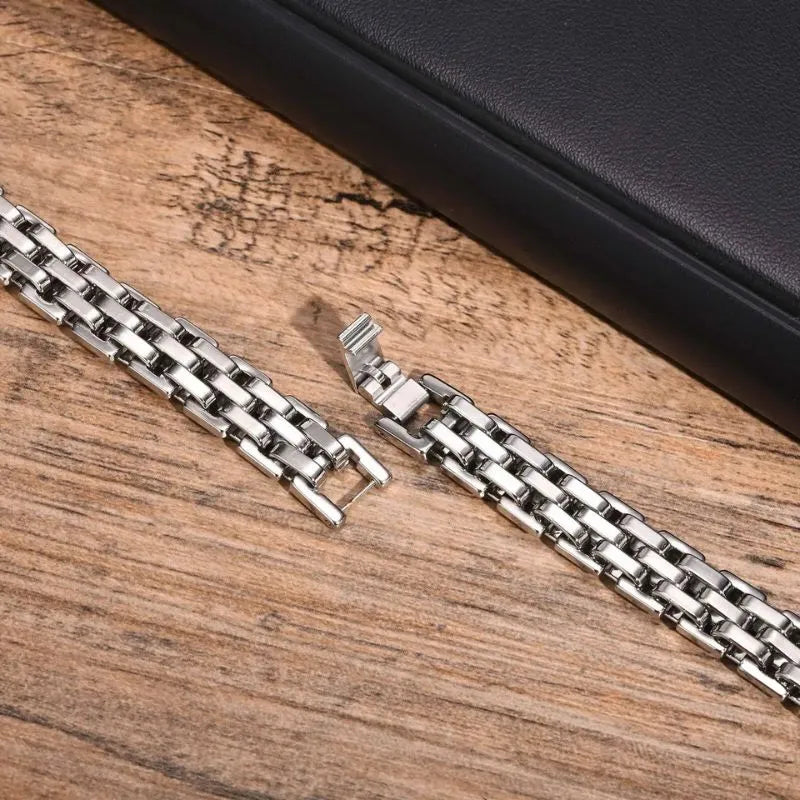 Silver watchband-link bracelet shown open and unclasped, laid diagonally on a wooden surface with a dark box prop.