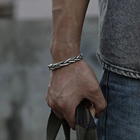 Silver twisted cuff bracelet worn on the wrist of a man holding a bag strap, wearing a denim jacket outdoors.
