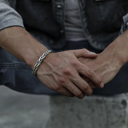 Silver twisted cuff bracelet worn on the wrist with hands clasped together, denim vest, close-up.