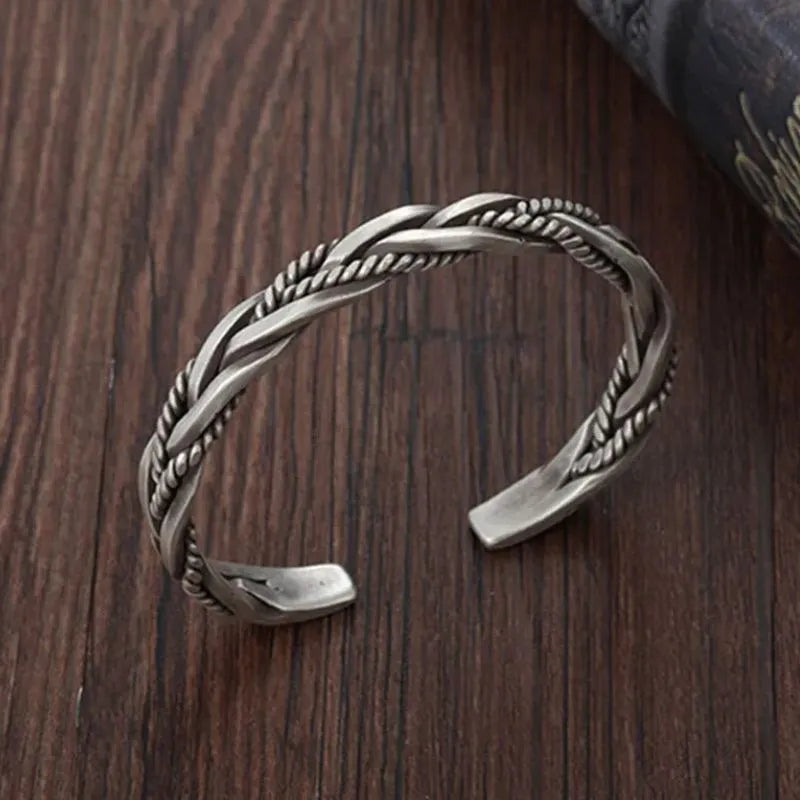 Silver open-cuff bracelet with a braided rope-and-flat-band twist pattern, laid on a wooden surface with a dark fabric prop.
