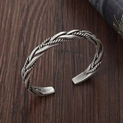 Silver open-cuff bracelet with a braided rope-and-flat-band twist pattern, laid on a wooden surface with a dark fabric prop.