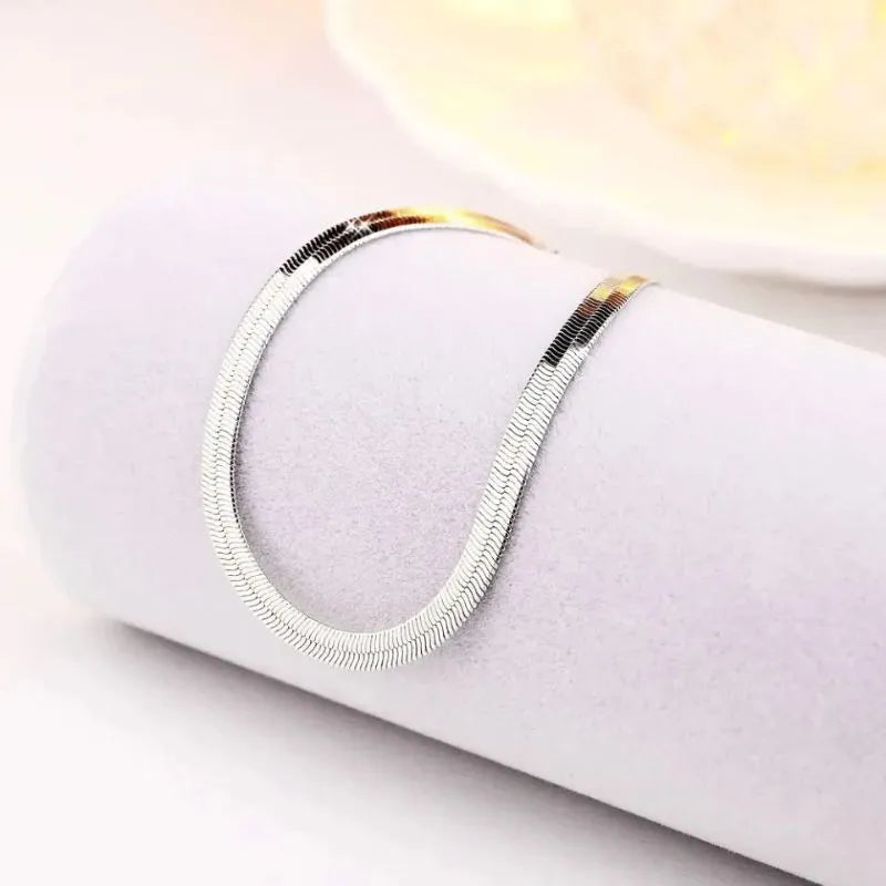 Silver flat blade chain bracelet draped over a light lavender cylindrical display stand, with warm golden light in the background.