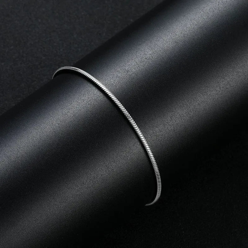 Silver thin snake chain bracelet draped over a black cylindrical display stand against a dark background.