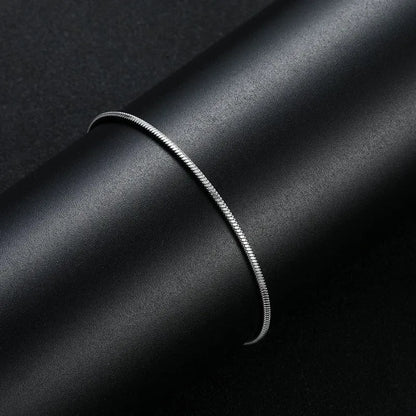 Silver thin snake chain bracelet draped over a black cylindrical display stand against a dark background.
