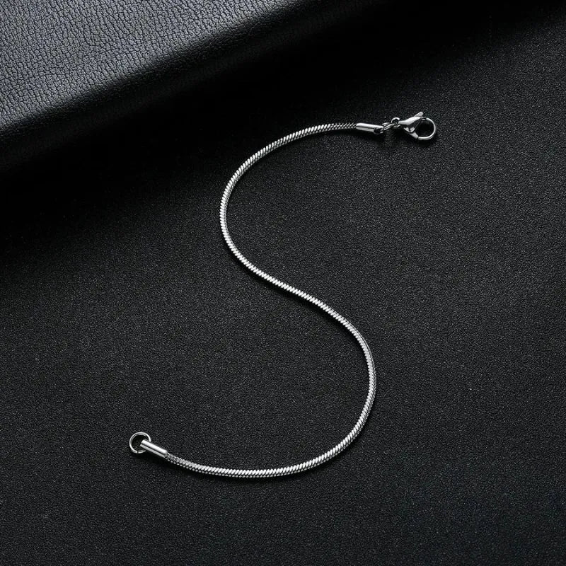 Silver snake chain bracelet laid in an S-curve on a black leather surface, with a dark leather book prop in the corner.