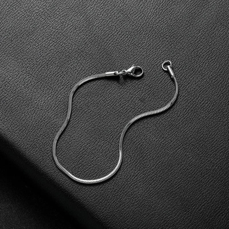 Silver thin snake chain bracelet shown open and unclasped, laid loosely on a black leather surface.