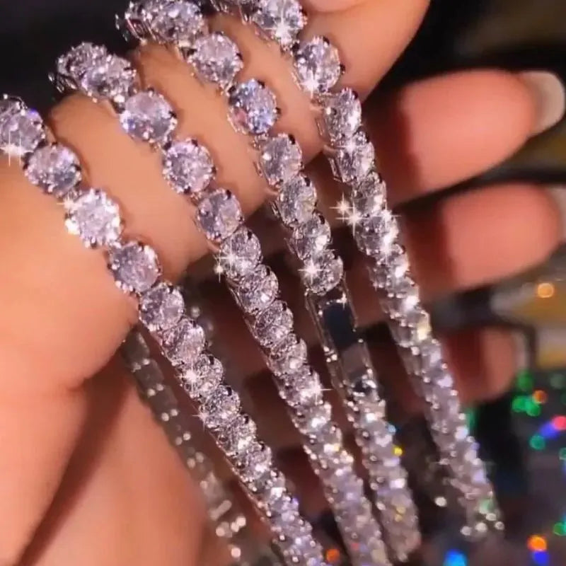 Multiple silver tennis bracelets held between fingers, sparkling under festive bokeh lighting.
