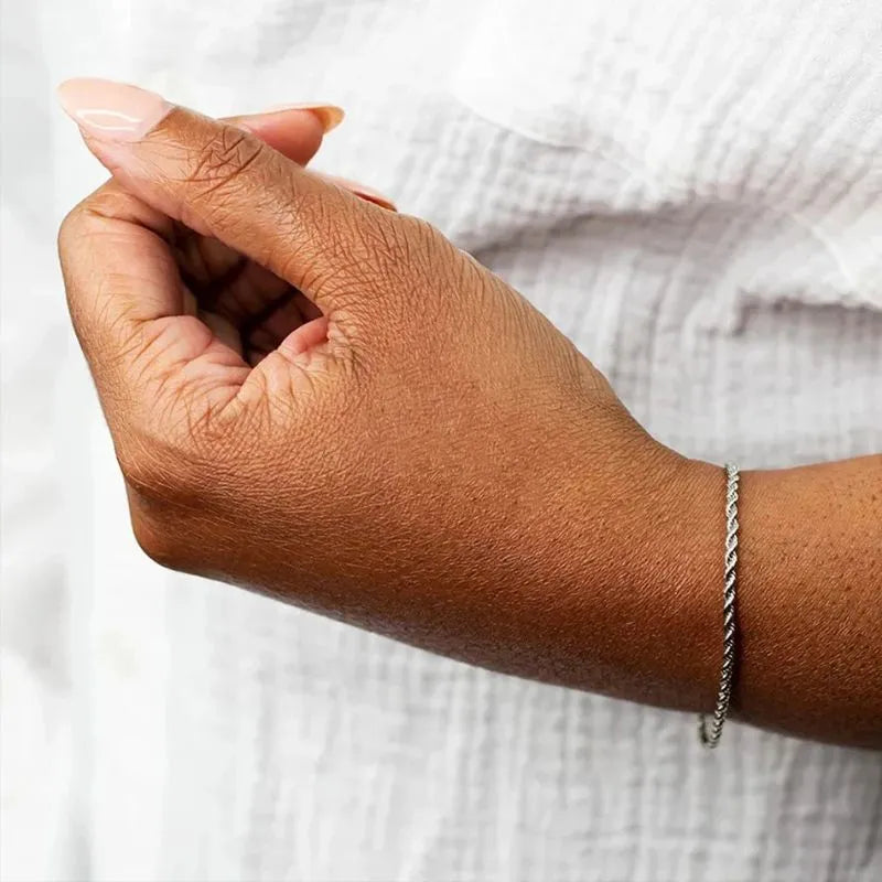 Silver twisted rope chain bracelet worn on the wrist of a relaxed hand near the chest, against a white textured clothing background.
