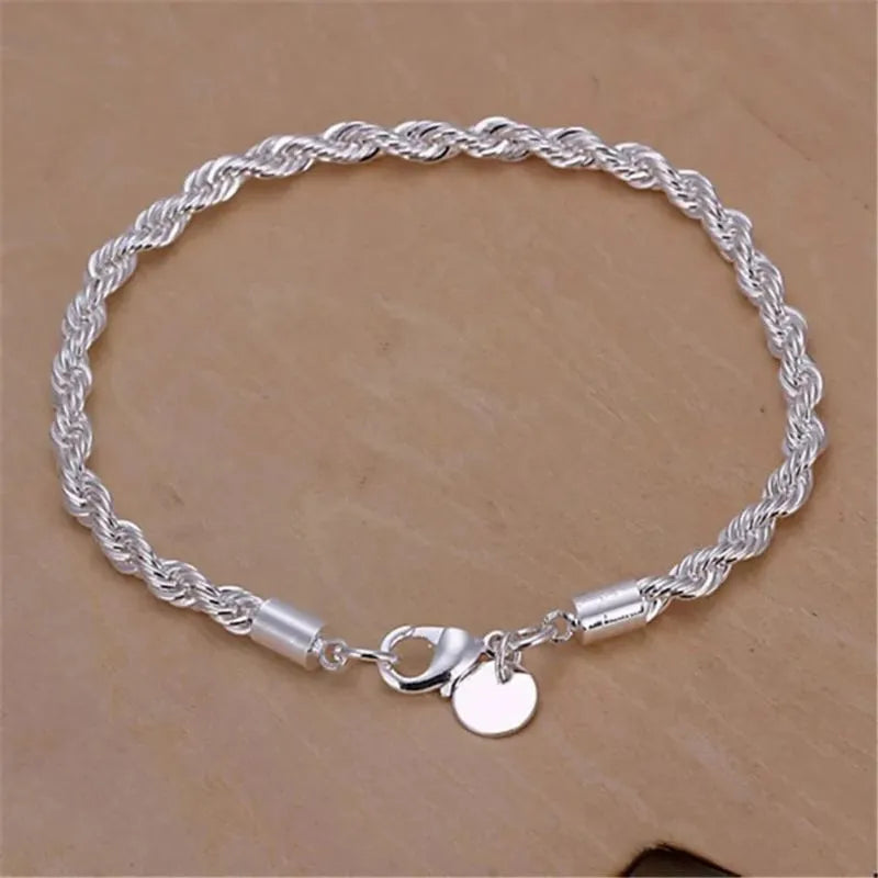 Silver twisted rope chain bracelet laid flat in an oval on a tan wooden surface, showing the lobster clasp and round tag charm.